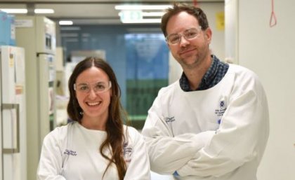 Two scientists wearing white lab coats, and goggles stand in a corridor smiling at the camera.